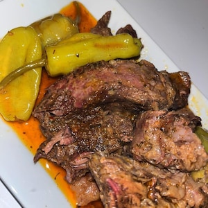May include: A serving of slow-cooked, shredded beef roast with a dark, seasoned crust and visible meat fibres, accompanied by two whole, pale yellow pickled pepperoncini peppers, all resting in a pool of reddish-brown savoury cooking juices.