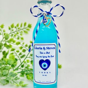 Może przedstawiać: Mała szklana butelka wypełniona niebieskim płynem, zwieńczona srebrną zakrętką. Ozdobiona niebiesko-białą kokardą ze sznurka i złotym sercem z okiem proroka. Etykieta głosi "Gloria & Steven" i "Vodka 50 ml."