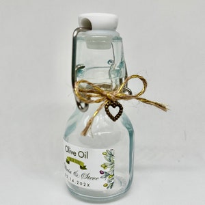Puede incluir: Una peque&ntilde;a botella de vidrio con una tapa blanca y un lazo de cuerda marr&oacute;n. La botella tiene una etiqueta que dice "Olive Oil with Love Olivia & Steve 02.14.202X".