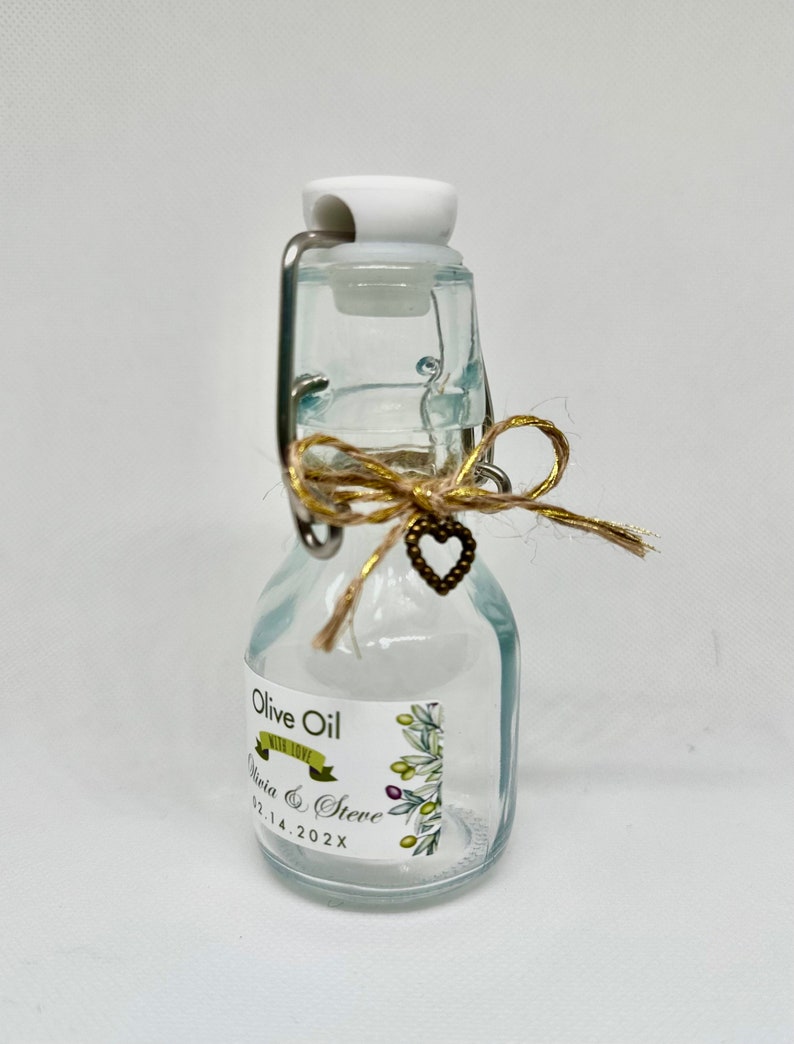 Puede incluir: Una peque&ntilde;a botella de vidrio con una tapa blanca y un lazo de cuerda marr&oacute;n. La botella tiene una etiqueta que dice "Olive Oil with Love Olivia & Steve 02.14.202X".