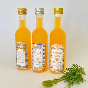 May include: Three small glass bottles filled with orange liquid and topped with gold and silver lids. Each bottle has a colorful label with a floral design and text that reads "Orangecello With Love" and a name, "Andrew's Christening", "Aida & Evan" and "Anika's Hen Party".