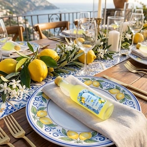 May include: A table setting with a coastal theme. A bottle of limoncello rests on a linen napkin atop a plate decorated with lemons. The table is set with gold cutlery, wine glasses, and fresh lemons. The background shows a scenic view of the ocean.