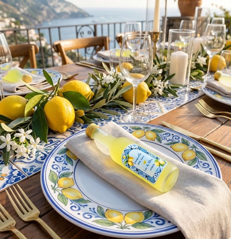 May include: A table setting with a coastal theme. A bottle of limoncello rests on a linen napkin atop a plate decorated with lemons. The table is set with gold cutlery, wine glasses, and fresh lemons. The background shows a scenic view of the ocean.