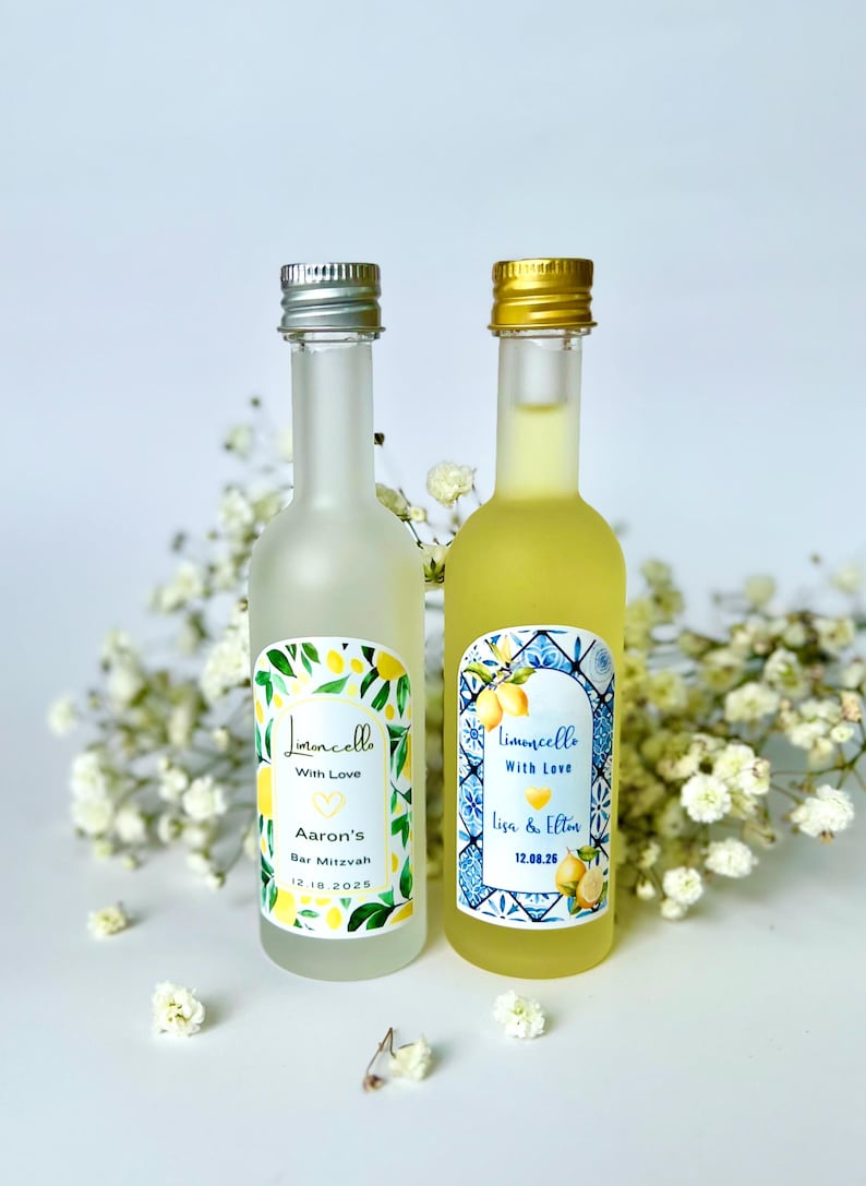 May include: Two small limoncello bottles with silver and gold caps. One bottle is frosted and the other is clear, both with labels featuring lemon illustrations and text. The bottles are surrounded by white flowers.