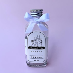Pode incluir: Garrafa de vidro transparente com tampa prateada e laço branco. O rótulo apresenta uma ilustração de casamento, nomes e a data 06.07.25. O rótulo também diz "SCOTCH" e "50 ml".