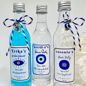 Może przedstawiać: Trzy małe szklane butelki z niebieskim płynem i etykietami. Etykiety mają wzór niebieskiego oka i tekst, który brzmi: "Erika's Bridal Shower", "Aretha's Hens Party" i "Antonia's Bach Party". Każda butelka ma niebieską i białą wstążkę zawiązaną wokół szyjki z niebieskim oczkiem.