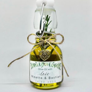 Puede incluir: Una peque&ntilde;a botella de vidrio llena de aceite de oliva y una ramita de romero. La botella tiene una tapa blanca con un anillo de metal y una etiqueta que dice "Olive Oil with love Ninette & Bastian 14.02.202X". Un peque&ntilde;o amuleto de coraz&oacute;n dorado cuelga de la botella.