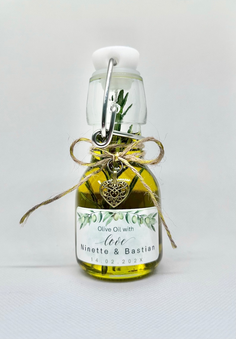 Puede incluir: Una peque&ntilde;a botella de vidrio llena de aceite de oliva y una ramita de romero. La botella tiene una tapa blanca con un anillo de metal y una etiqueta que dice "Olive Oil with love Ninette & Bastian 14.02.202X". Un peque&ntilde;o amuleto de coraz&oacute;n dorado cuelga de la botella.