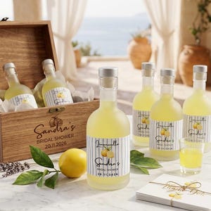 May include: A wooden crate containing glass bottles filled with yellow liquid, each with a label reading Limoncello, Sandra's Bridal Shower, 20.11.2026. A fresh lemon, green leaves, a small glass of yellow liquid, and a stack of thank you cards are nearby.