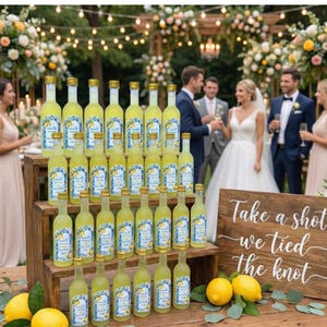 May include: A display of small glass bottles filled with yellow liquid, arranged on wooden crates. Each bottle has a gold cap and a label with a blue and white design. The bottles are part of a wedding reception, with a sign that says "Take a shot we tied the knot."