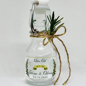 Puede incluir: Una peque&ntilde;a botella de vidrio con un tap&oacute;n de corcho y una etiqueta que dice "Olive Oil with Love Gloria & Oliver 02.14.202X". La botella est&aacute; decorada con una ramita de romero y un lazo de cuerda.