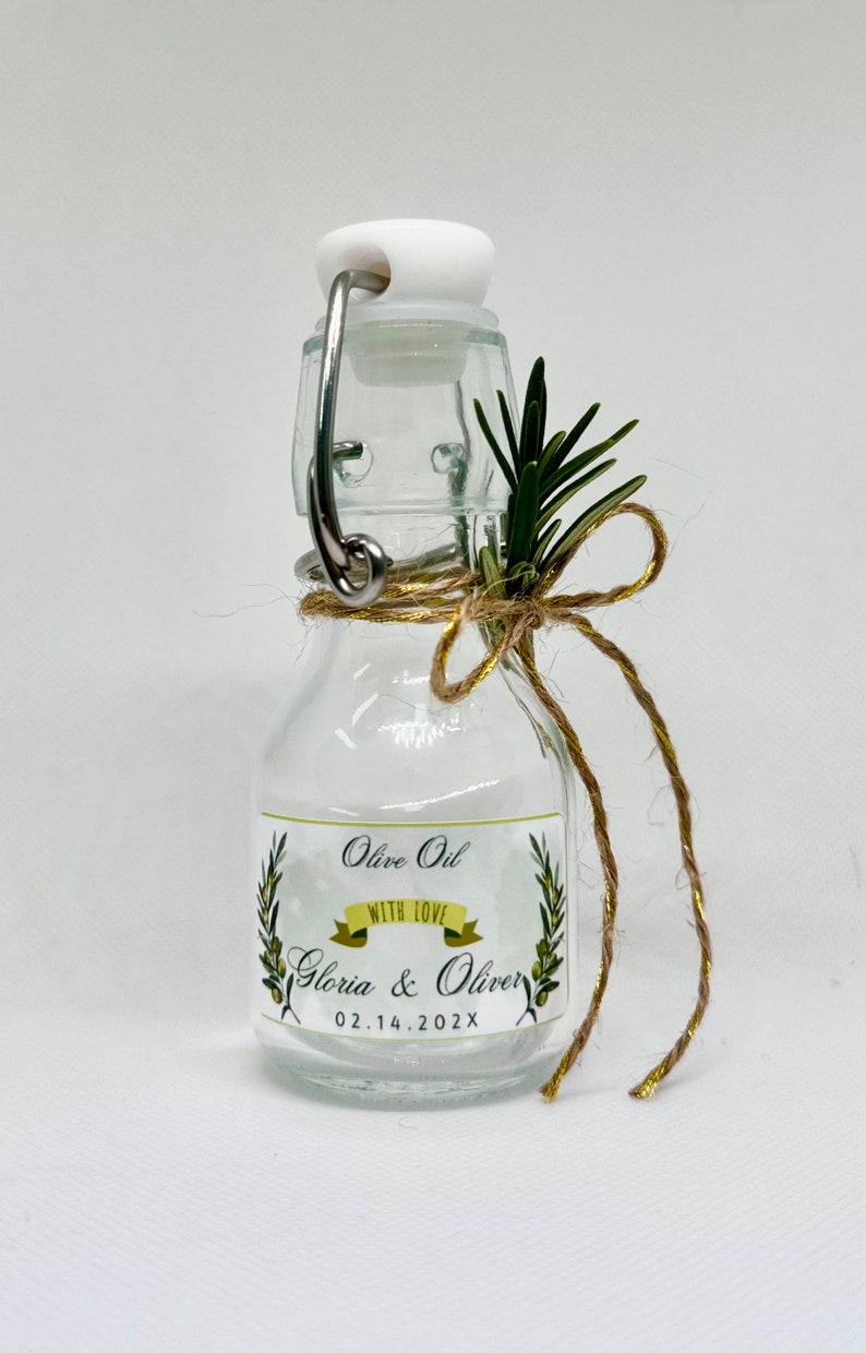 Puede incluir: Una peque&ntilde;a botella de vidrio con un tap&oacute;n de corcho y una etiqueta que dice "Olive Oil with Love Gloria & Oliver 02.14.202X". La botella est&aacute; decorada con una ramita de romero y un lazo de cuerda.