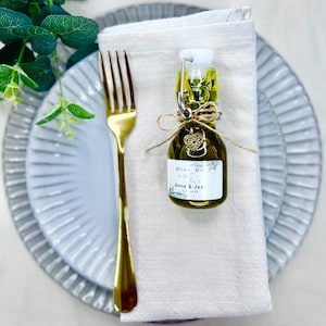 Puede incluir: Una peque&ntilde;a botella de vidrio de aceite de oliva con una etiqueta blanca que dice "Olive Oil Favor Anne & Jes 12.0.2022". La botella est&aacute; atada con un cordel y un peque&ntilde;o dije de coraz&oacute;n dorado. La botella est&aacute; sentada sobre una servilleta de lino blanca en un plato gris.