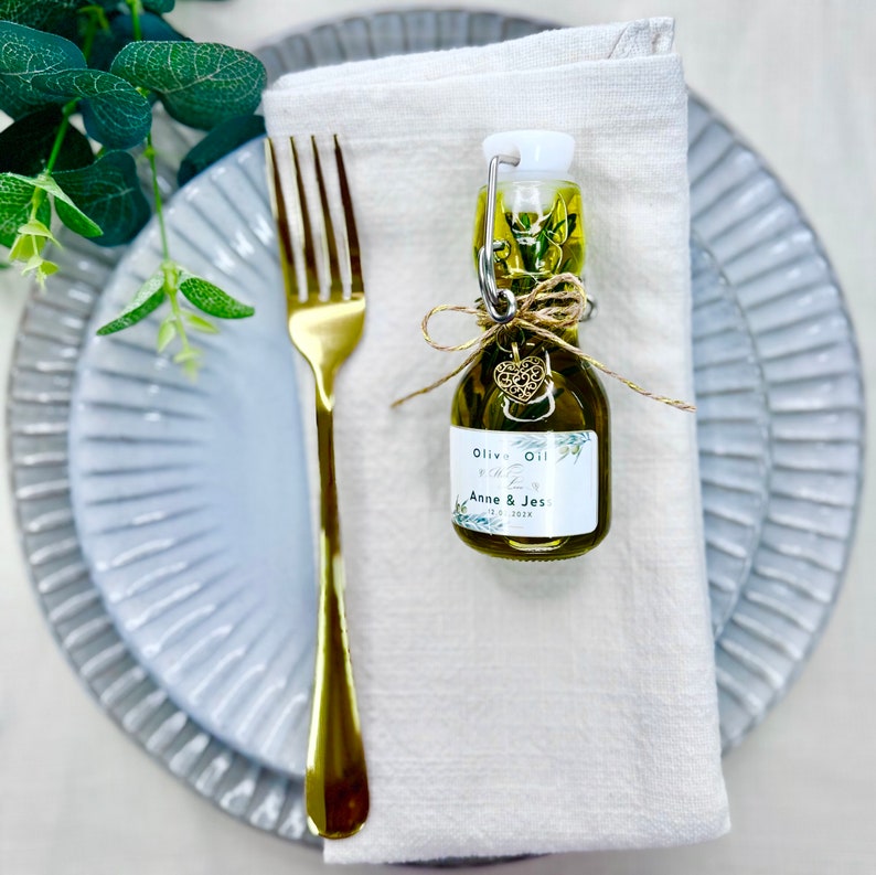 Puede incluir: Una peque&ntilde;a botella de vidrio de aceite de oliva con una etiqueta blanca que dice "Olive Oil Favor Anne & Jes 12.0.2022". La botella est&aacute; atada con un cordel y un peque&ntilde;o dije de coraz&oacute;n dorado. La botella est&aacute; sentada sobre una servilleta de lino blanca en un plato gris.