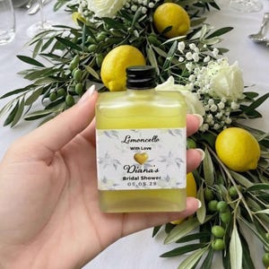 May include: A small, square bottle of limoncello liqueur with a black cap, held in a hand. The label reads "Limoncello With Love" and "Diana's Bridal Shower 05.05.28". The bottle is surrounded by lemons, white flowers, and greenery.