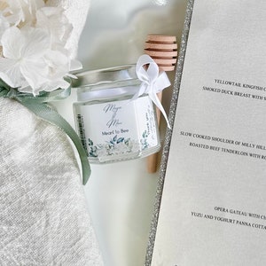 Peut inclure: Un petit pot en verre avec une étiquette blanche sur laquelle est inscrit "May & Ma Meant to Bee". Le pot est attaché avec un ruban blanc et un bâtonnet de miel en bois se trouve à côté.