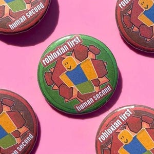 May include: A collection of round buttons with a Roblox theme. The central button is green with the text "robloxian first" and "human second" surrounding a Roblox character. Other buttons are red and brown with the same text.