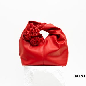 May include: A red handbag with three rose embellishments. The bag is made of a smooth material and has a handle. The word "MINI" is printed in black on the bottom right.
