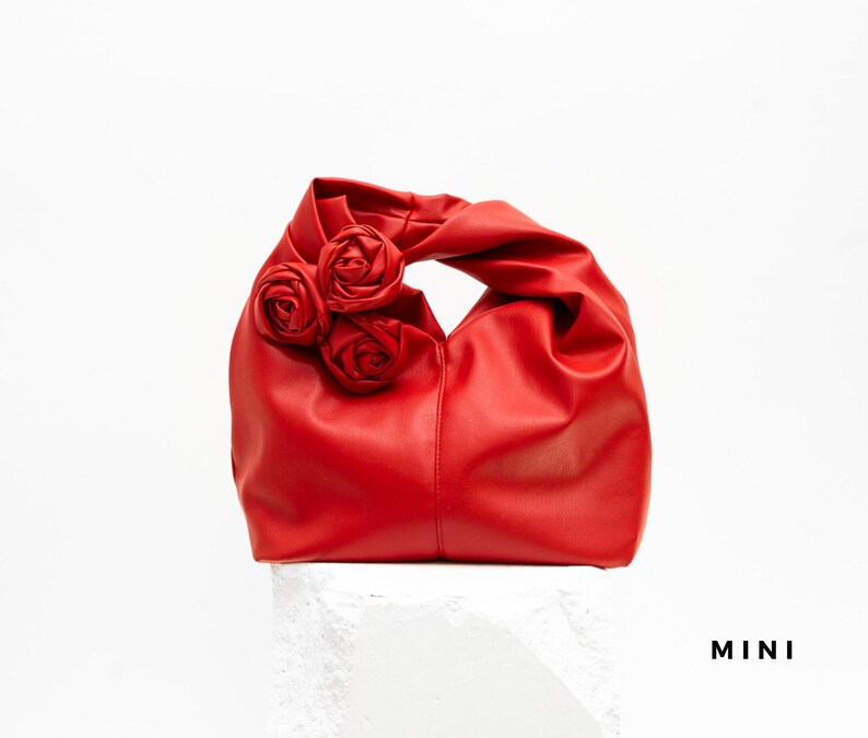 May include: A red handbag with three rose embellishments. The bag is made of a smooth material and has a handle. The word "MINI" is printed in black on the bottom right.