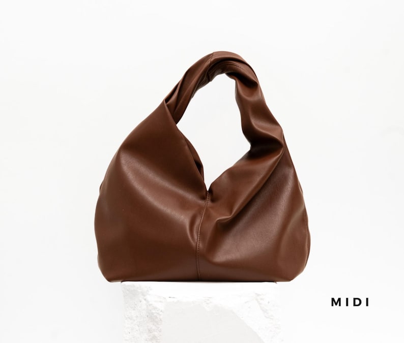 May include: A brown leather handbag with a twisted handle. The bag has a soft, slouchy design and is displayed against a white background. The word "MIDI" is printed in black on the bottom right.