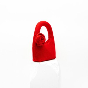 May include: A small, bright red handbag with a rounded handle and a fabric rose detail. The bag is made of a felt-like material and sits on a white surface. The overall design is simple and elegant.