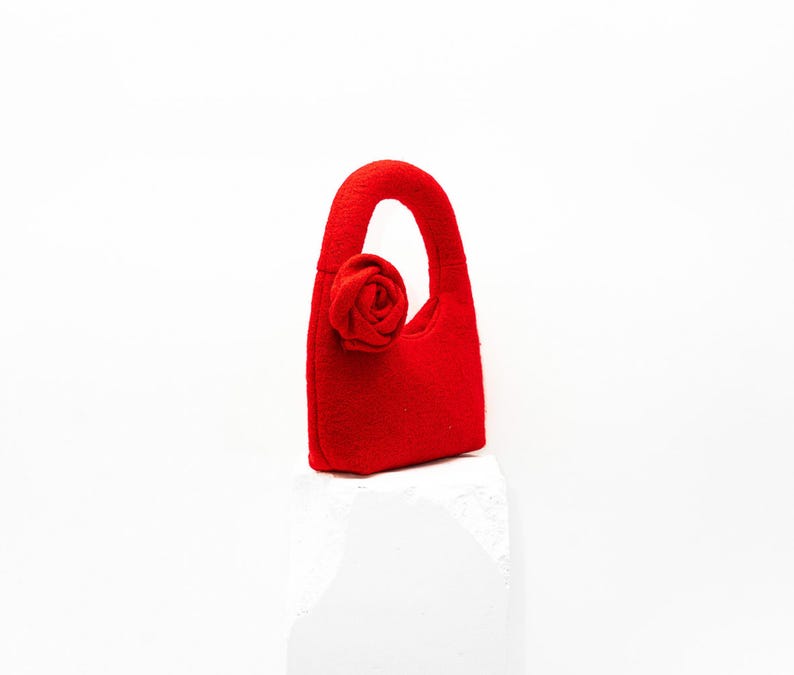 May include: A small, bright red handbag with a rounded handle and a fabric rose detail. The bag is made of a felt-like material and sits on a white surface. The overall design is simple and elegant.