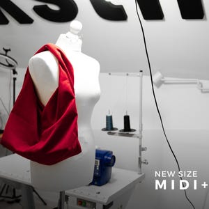 May include: A red fabric bag draped over a white mannequin. The bag is a solid, vibrant red. The background includes a sewing machine, spools of thread, and the text "NEW SIZE MIDI+".