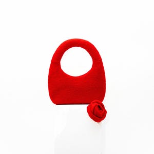 May include: A vibrant red handbag with a rounded handle and a matching red rose. The bag has a smooth texture and a simple, elegant design, perfect for carrying essentials. The rose is a small, decorative accent.