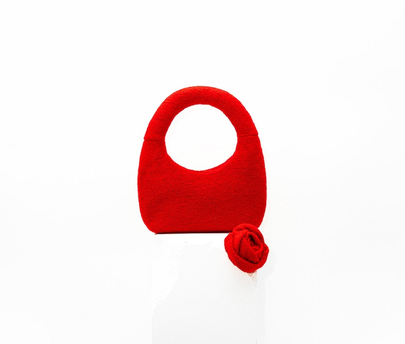 May include: A vibrant red handbag with a rounded handle and a matching red rose. The bag has a smooth texture and a simple, elegant design, perfect for carrying essentials. The rose is a small, decorative accent.