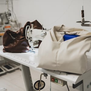 May include: A white sewing machine sits on a white table, with a brown leather bag and a cream-colored bag. A blue box is visible inside the cream bag. Spools of thread are mounted above the table. The scene suggests a leather goods workshop.