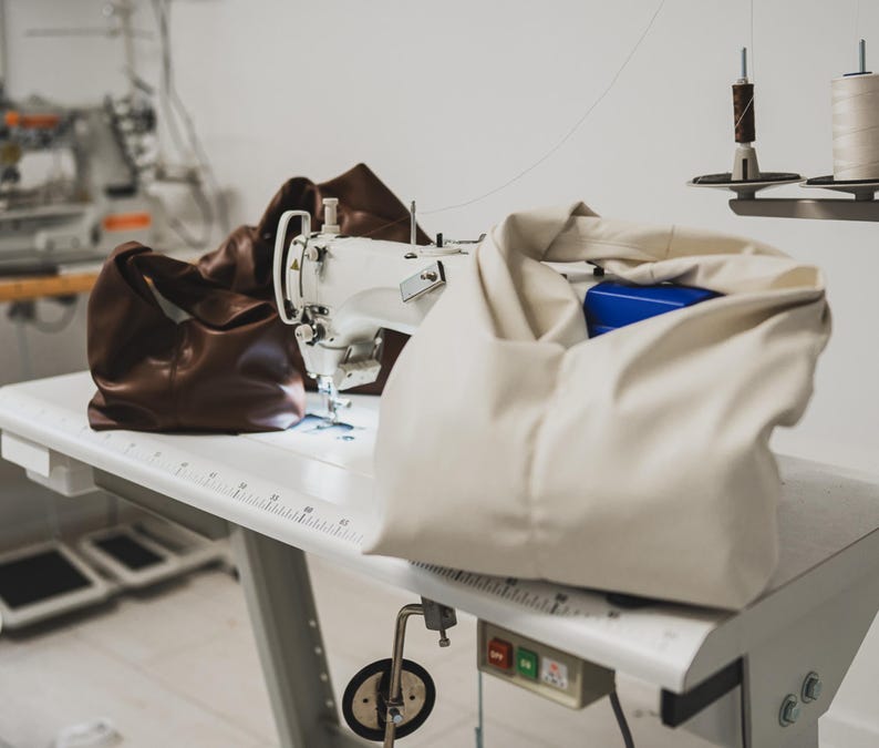 May include: A white sewing machine sits on a white table, with a brown leather bag and a cream-colored bag. A blue box is visible inside the cream bag. Spools of thread are mounted above the table. The scene suggests a leather goods workshop.
