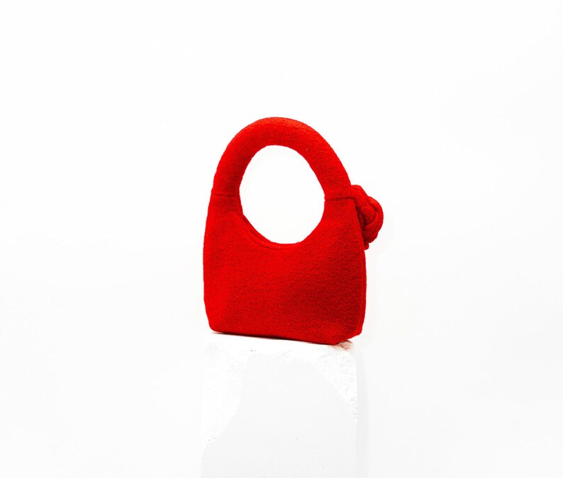 May include: A small, bright red handbag with a rounded handle and a decorative knot on the side. The bag is made of a soft material and has a simple, elegant design. The bag is suitable for carrying small items.