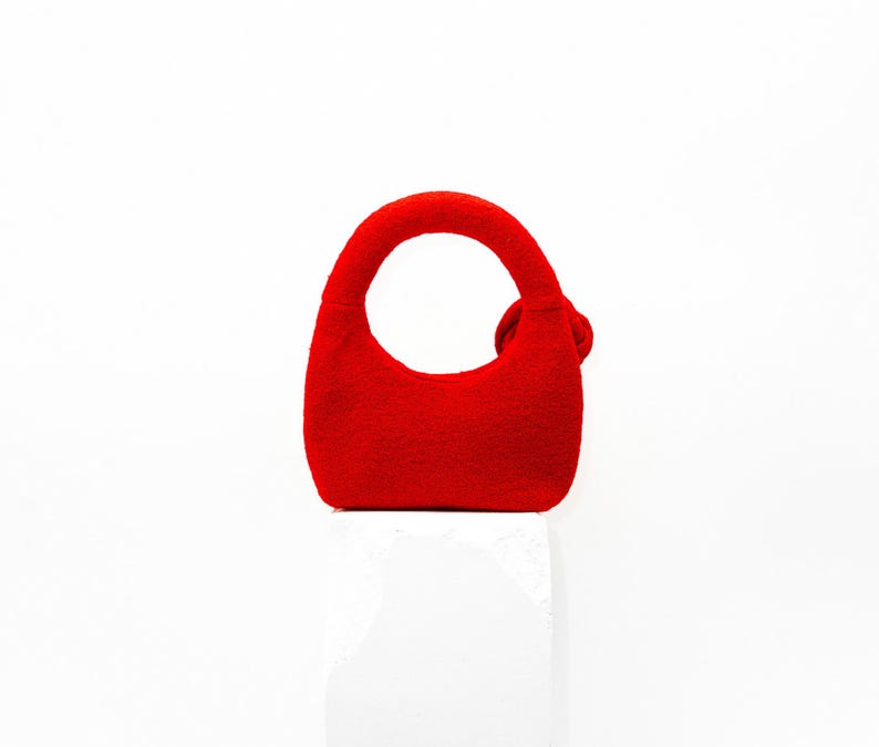 May include: A small, bright red handbag with a rounded handle and a small decorative accent. The bag is made of a textured material and sits on a white surface. The bag's design is simple and modern.