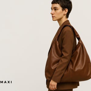May include: A large, brown leather shoulder bag with a soft, slouchy design. The bag is carried over the shoulder and has a wide strap. The word "MAXI" is printed in black at the bottom left corner of the image. The bag is paired with a brown blazer.
