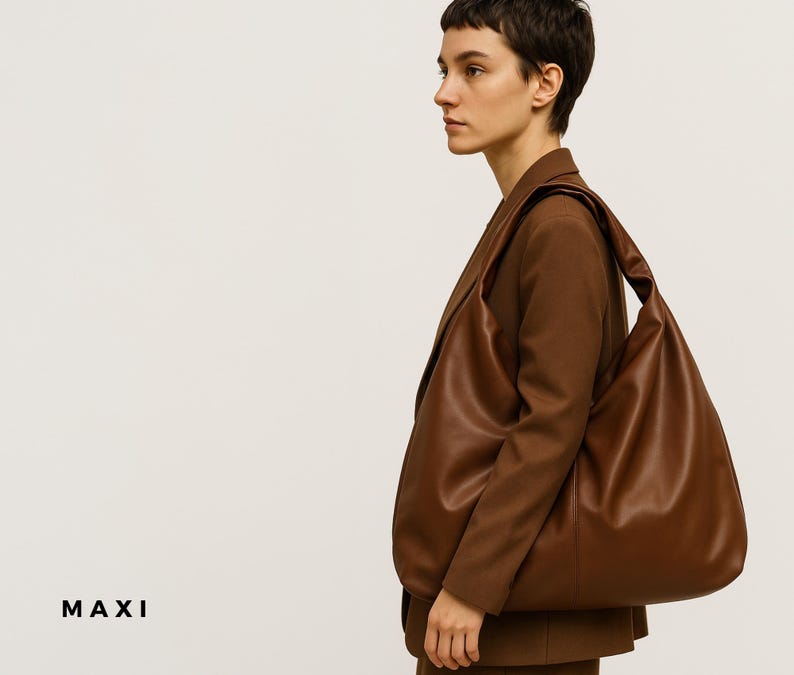 May include: A large, brown leather shoulder bag with a soft, slouchy design. The bag is carried over the shoulder and has a wide strap. The word "MAXI" is printed in black at the bottom left corner of the image. The bag is paired with a brown blazer.