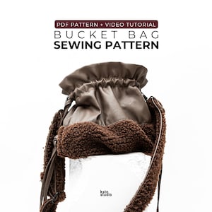 May include: A brown bucket bag sewing pattern with a video tutorial. The bag is made of brown leather-like material with a fluffy, brown trim. The bag has a drawstring closure and long straps. The text "PDF PATTERN + VIDEO TUTORIAL BUCKET BAG SEWING PATTERN" is at the top.
