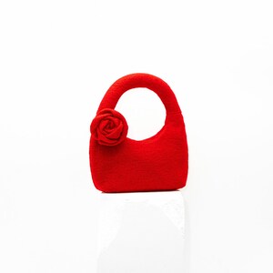 May include: A small, bright red handbag with a rounded handle and a decorative rose on the side. The bag is made of a textured material and sits on a white surface. The bag's design is simple and elegant.