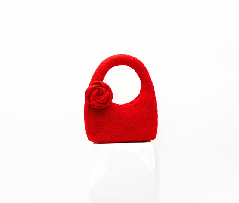 May include: A small, bright red handbag with a rounded handle and a decorative rose on the side. The bag is made of a textured material and sits on a white surface. The bag's design is simple and elegant.