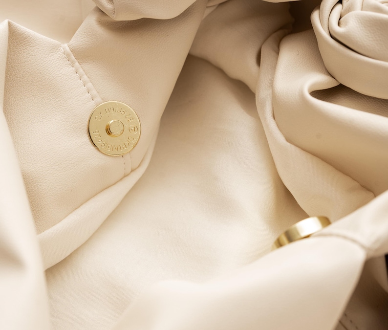 May include: Close-up of a cream-colored handbag with a gold magnetic clasp. The bag's soft, folded material creates a textured appearance. A gold ring is also visible within the bag's interior.