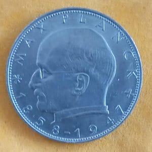 Könnte beinhalten: Eine silberne Münze mit dem Profil eines Mannes mit Brille. Der Rand der Münze trägt Text, darunter das Jahr 1941 und das Wort "PLANA". Die Münze liegt auf einem gelben Hintergrund.