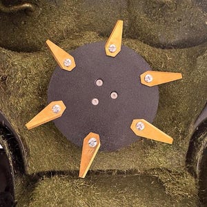 Könnte beinhalten: Nahaufnahme einer Rasenmäher-Messeranordnung. Eine schwarze kreisförmige Klinge mit acht befestigten, hellbraunen, abgewinkelten Klingen. Die Klingen sind mit silbernen Schrauben befestigt. Die Anordnung ist von grünem Gras umgeben.