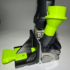 Può includere: Un accessorio per utensili elettrici stampato in 3D di colore nero e verde. L'accessorio ha una leva verde e un imbuto di raccolta della polvere verde. Lo strumento è montato su una base nera.