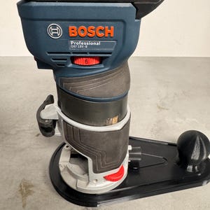 Könnte beinhalten: Ein kabelloser Bosch Professional GKF 18V-8 Fräser. Das Werkzeug ist hauptsächlich blau und schwarz, mit roten Akzenten an den Einstellrädern. Das Bosch-Logo ist deutlich sichtbar. Der Fräser steht auf einer schwarzen Basis.