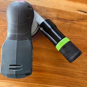 Op de afbeelding: Een zwart en groen elektrisch gereedschap met een schuurpad en een lange, zwarte handgreep. Het gereedschap heeft een groen label met de tekst "TOOL" erop.