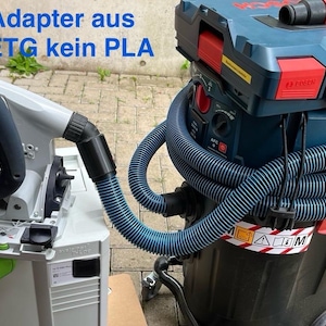 Op de afbeelding: Een groen en zwart elektrisch gereedschap met een zwarte slang die is aangesloten op een blauwe en zwarte stofzuiger. De stofzuiger heeft een rood Bosch-logo en een waarschuwingssticker met een gele driehoek en een uitroepteken. De tekst op het elektrische gereedschap luidt "Adapter aus PETG kein PLA".