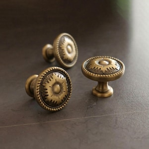 Puede incluir: Tres tiradores de armario de latón antiguo con un diseño detallado de sol. Los tiradores tienen una cara redonda con un borde en forma de cuerda y un diseño circular central. Los tiradores están sobre un fondo gris oscuro.
