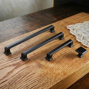 Könnte beinhalten: Ein Satz von vier schwarzen Schrankbeschlägen, darunter drei rechteckige Griffe und ein quadratischer Knopf. Die Griffe haben ein gerilltes Design und werden auf einer Holzoberfläche präsentiert. Die Beschläge sind wahrscheinlich für Schränke oder Schubladen.