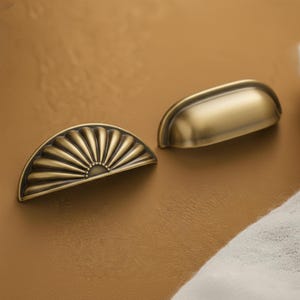 Könnte beinhalten: Zwei antike Messing-Schrankgriffe. Einer ist ein Halbkreis mit einem fächerartigen Design, der andere ein einfacher, gebogener Muschelgriff. Beide haben eine warme, gealterte Oberfläche, die einen Vintage- oder Retro-Stil suggeriert.