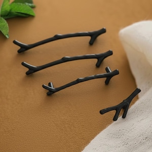 May include: Four black twig-shaped cabinet pulls. The pulls are made of metal and have a matte finish. The pulls are arranged on a brown surface with a white cloth in the background. The pulls are designed to look like small tree branches.