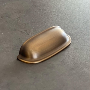 Könnte beinhalten: Ein bronzefarbener, schalenförmiger Schrankgriff mit geschwungenem Design. Die Hardware hat eine glatte, polierte Oberfläche und ist für die Montage an einer Schublade oder Schranktür konzipiert. Der Griff ist oval geformt und hat eine leichte Lippe am oberen Rand.